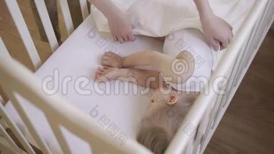 两岁的小女孩睡在铺着白色毯子的婴儿床上。 妈妈给宝宝盖上毯子.. 日间睡眠